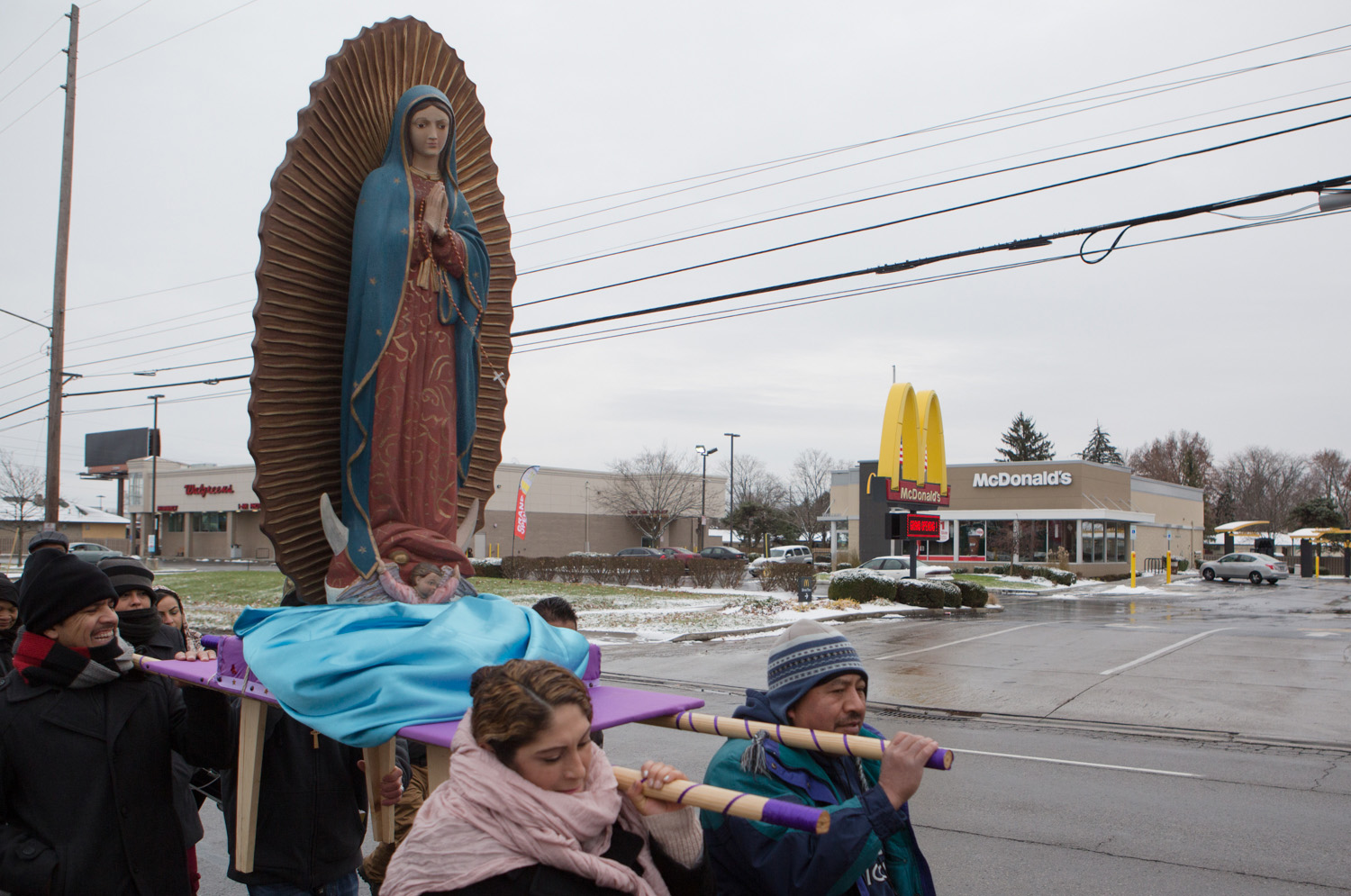 Crowd of people carrying a statue of Our Lady of Guadalupe past a McDonald's on a gray winter day