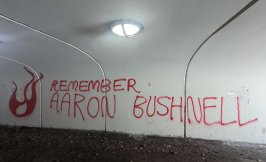 "Remember Aaron Bushnell," written in red spray paint next to a spray painted flame illustration, in a white-walled underground tunnel.