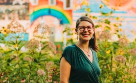 Headshot of a woman in front of an uban garden and outdoor mural. Woman has short, brown hair, glasses, and is wearing an emerald green shirt