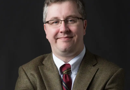 Headshot of a man with dark gray hair and glasses, light skin, wearing a tweed blazer and maroon tie. 