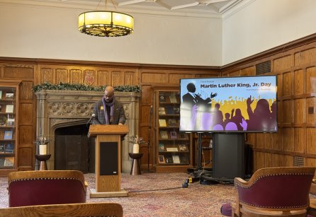 A man with dark skin stands behind a podium in a wood-paneled room. 