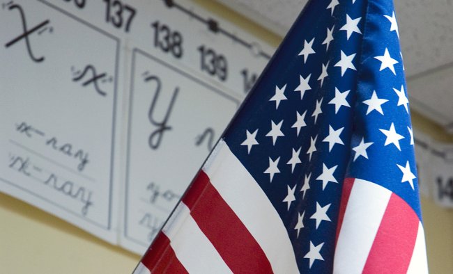 An American flag hangs in a classroom with the English alphabet on the wall behind it.