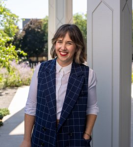 Woman with short, dark hair wearing a navy blue vest leans against a column outside a building. 
