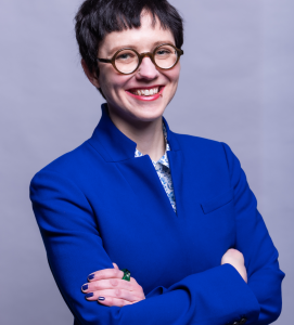 Headshot of a person with short dark hair and glasses in a cobalt blue blazer.