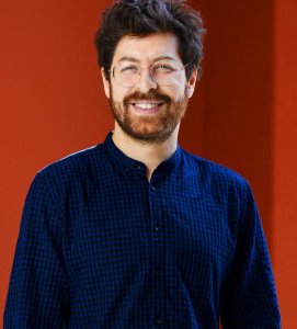 Headshot of a man with a red beard and glasses in front of an orange wall. 