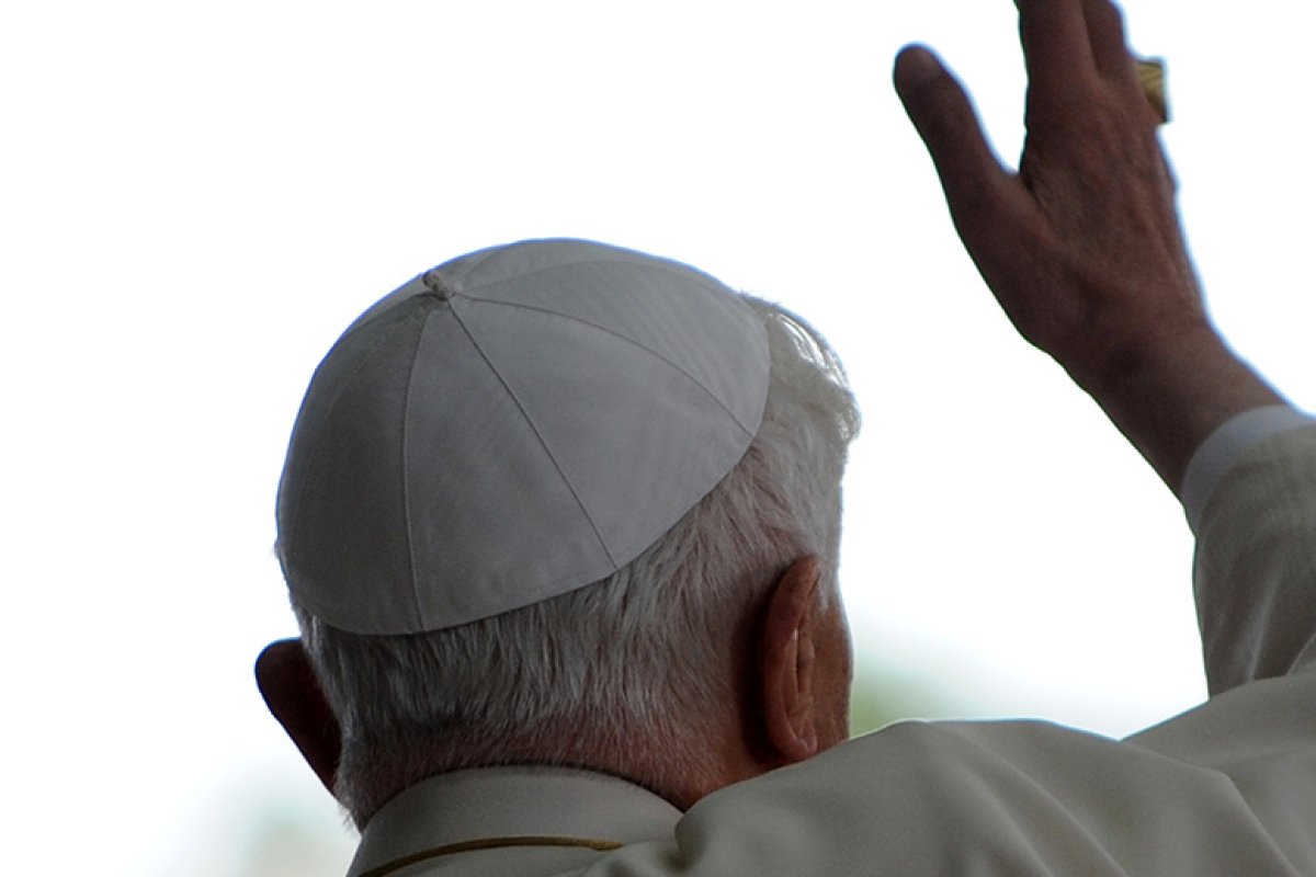 The Pope in a white hat and white robe waves with his right hand with his back turned toward the camera.