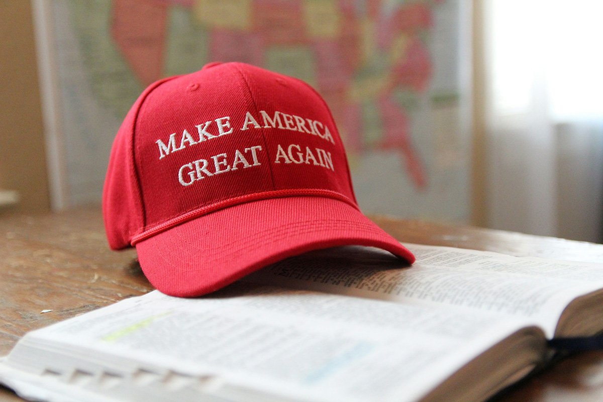 Red "Make America Great Again" baseball cap rests on an open Bible; a blurry map of the United States is visible in the background.