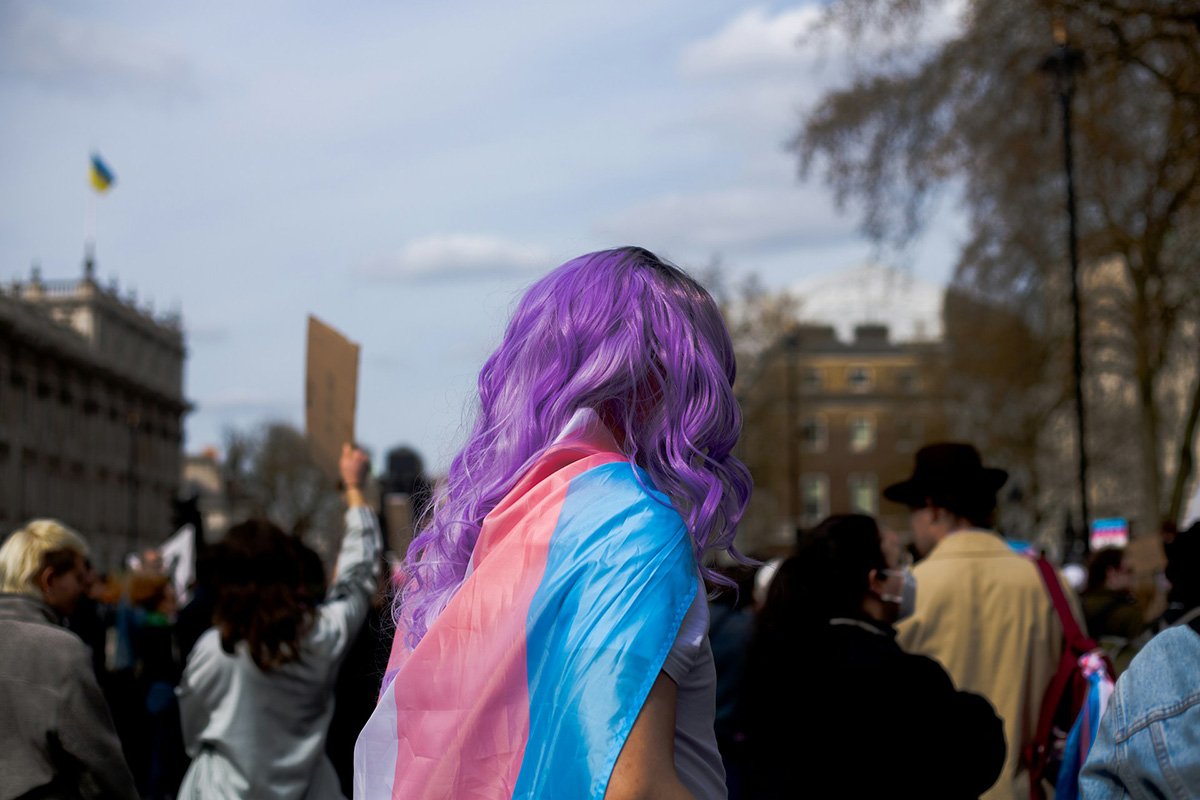 A person with long purple hair wears the pink and blue transgender flag in the style of a cape.