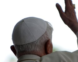 The Pope in a white hat and white robe waves with his right hand with his back turned toward the camera.