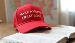 Red "Make America Great Again" baseball cap rests on an open Bible; a blurry map of the United States is visible in the background.