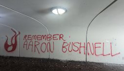 "Remember Aaron Bushnell," written in red spray paint next to a spray painted flame illustration, in a white-walled underground tunnel.